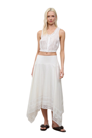 A blonde model wearing a white two-piece outfit consisting of a sleeveless lace-trimmed crop top and a high-low skirt, paired with dark brown slide sandals, against a plain white background.