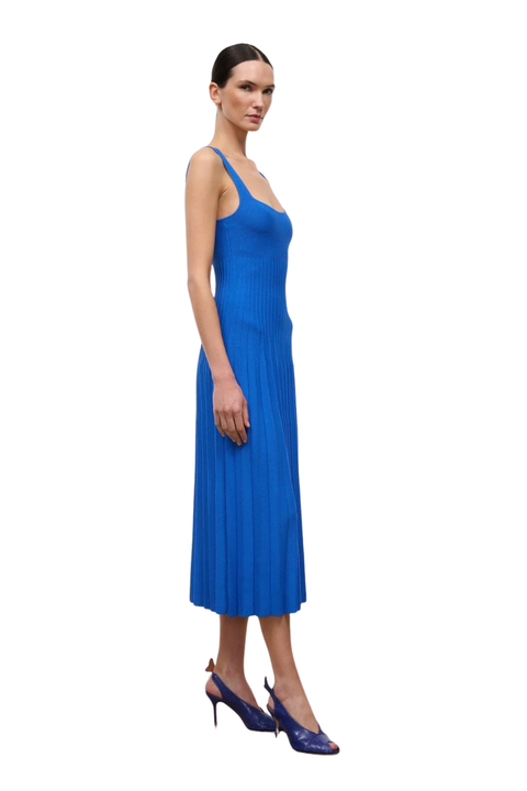A woman modeling a sleeveless, pleated midi dress in a vibrant blue, paired with dark blue patent high heels, set against a plain white background.
