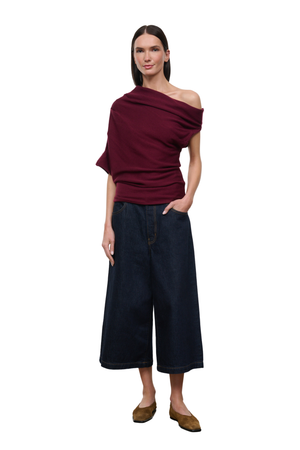 A woman modeling a dark burgundy draped off-the-shoulder top and dark wide-leg denim pants against a plain white background.