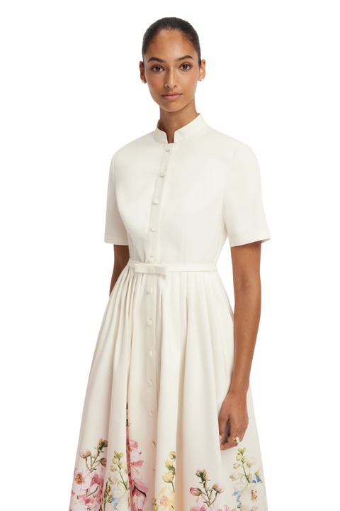 A woman modeling a short-sleeve midi dress with a stand collar, button front, and a soft floral print concentrated along the lower skirt, photographed against a plain white studio background.