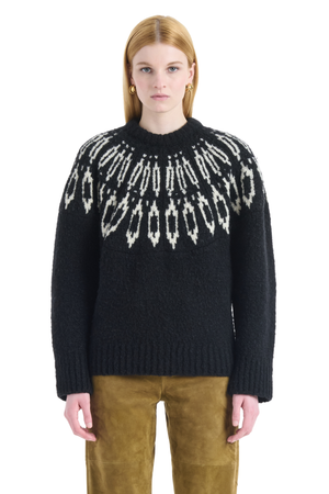 A model wearing a black knit sweater with a white geometric Fair Isle pattern across the chest, paired with brown pants, against a plain white background.