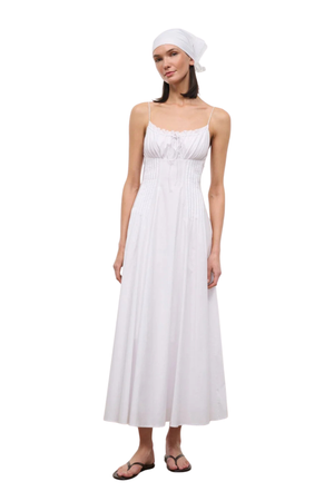A woman modeling a white sleeveless maxi dress with thin straps, a smocked bodice, and a flowing skirt, paired with a matching white headscarf and dark sandals, against a plain white background.
