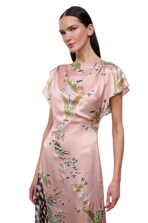 A woman modeling a light pink satin dress with a green and gold bird and floral print, set against a plain white background.
