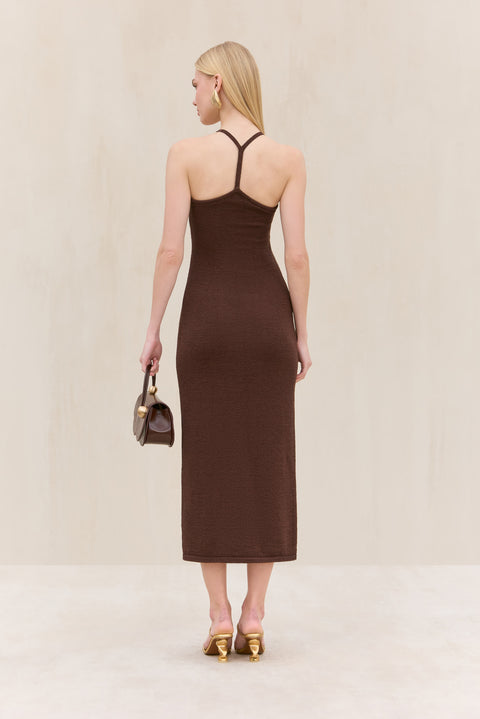 Back-facing woman wearing a brown knit midi dress that features a thin racerback strap, paired with a brown handbag and gold heels.