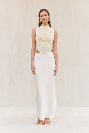 Woman wearing a cropped white tank top that features rows of pearl-beaded strands draped across the bodice, paired with a white maxi skirt.
