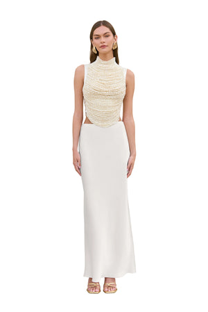Woman wearing a cropped white tank top that features rows of pearl-beaded strands draped across the bodice, paired with a white maxi skirt.
