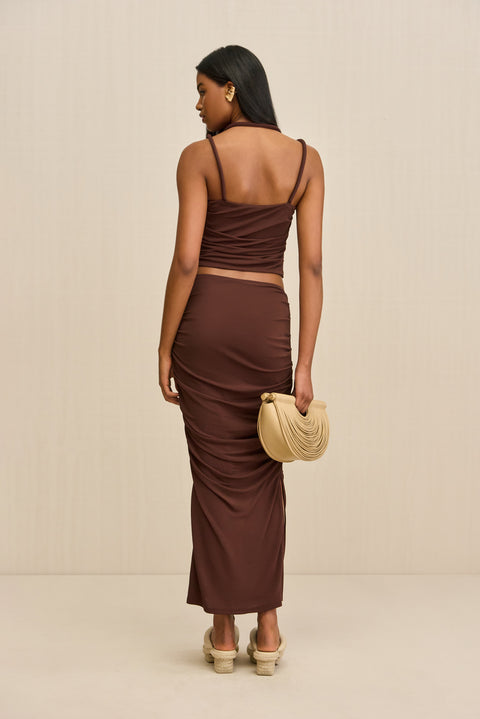 back-facing woman wearing a brown tank top that features a knotted double strap detail and a matching brown maxi skirt, paired with tan sandals and handbag.
