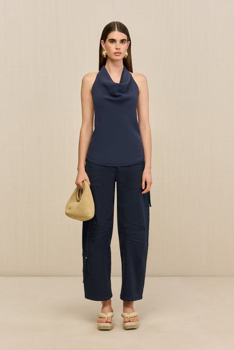 Front-facing woman wearing a navy blue sleeveless top that features a cowl halter neck, paired with blue pants, natural sandals, and a woven handbag.
