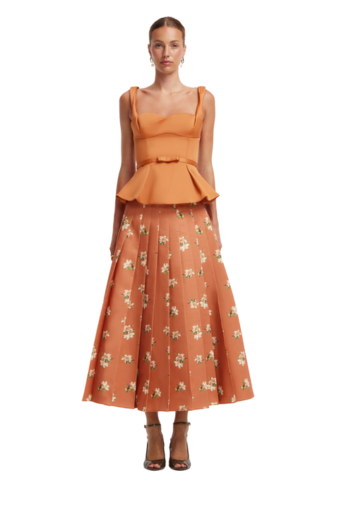 A model wearing a two-piece orange set with a sleeveless peplum top and a full skirt featuring a small white floral pattern, paired with brown strappy heels against a plain white background.