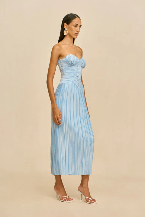 Woman wearing a light blue strapless pleated dress against a beige background standing to the side.