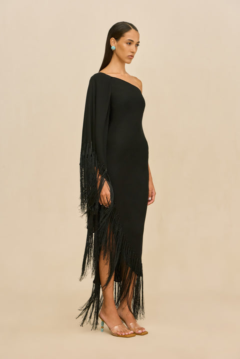 Woman wearing a black one-shoulder dress with fringe details on a beige background facing the side.