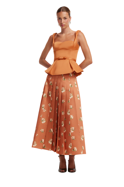 A woman modeling a sleeveless orange peplum top paired with a matching orange pleated midi skirt featuring a small white floral print, set against a plain white background.