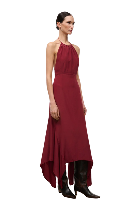 A woman in a sleeveless halter-neck maxi dress in deep burgundy, styled with black knee-high boots against a plain white background.