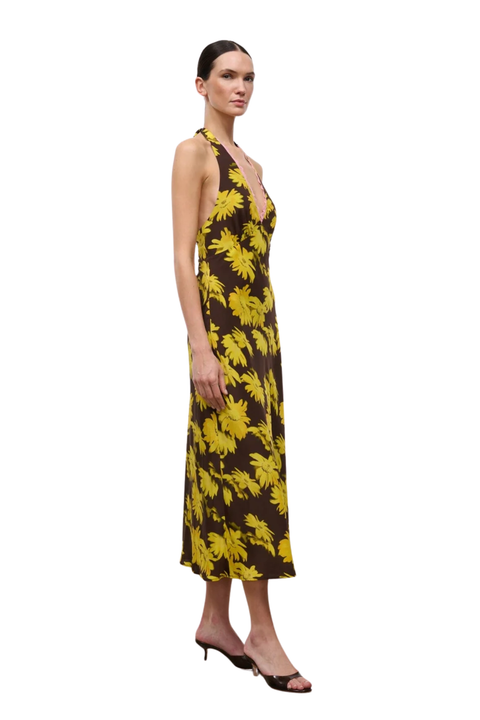 A woman modeling a sleeveless halter-neck midi dress with a bold yellow tropical leaf print on a dark brown background, paired with black heeled sandals, set against a plain white studio background.