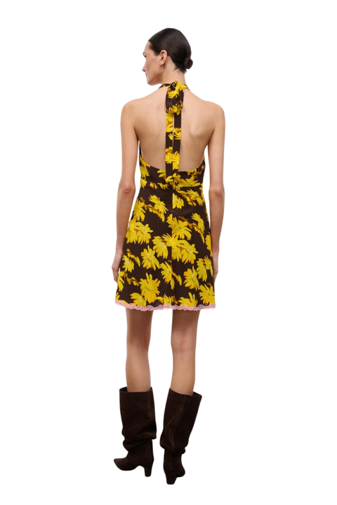 A model shown from the back wearing a halter-neck mini dress with a yellow and brown floral pattern, paired with dark brown knee-high boots, against a plain white background.