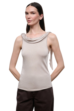 A woman modeling a sleeveless champagne colored top with a draped cowl neckline and a side tie detail, paired with dark brown pants.