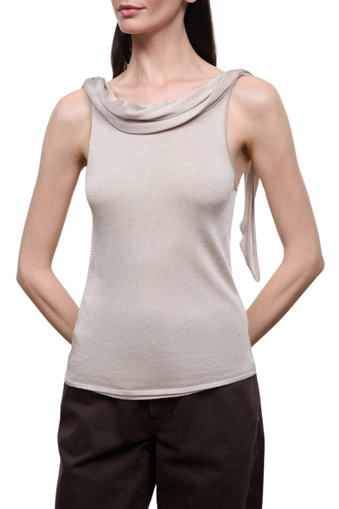 A woman modeling a sleeveless champagne colored top with a draped cowl neckline and a side tie detail, paired with dark brown pants.