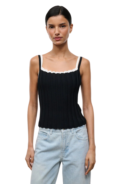 A woman modeling a black ribbed knit camisole top with a white ruffled trim along the scoop neckline, paired with light-wash jeans, against a plain white background.
