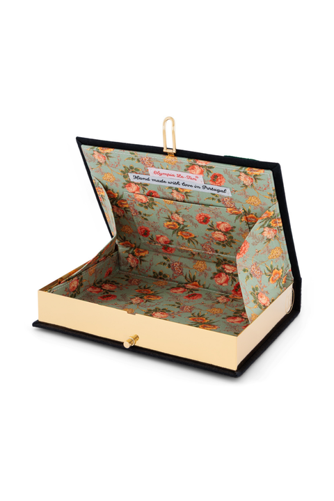 A book-shaped clutch purse designed to look like an open hardcover book, featuring a black cover, cream pages, and a floral patterned lining in sage green, orange, and pink. A gold-tone paperclip is clipped to the top edge.