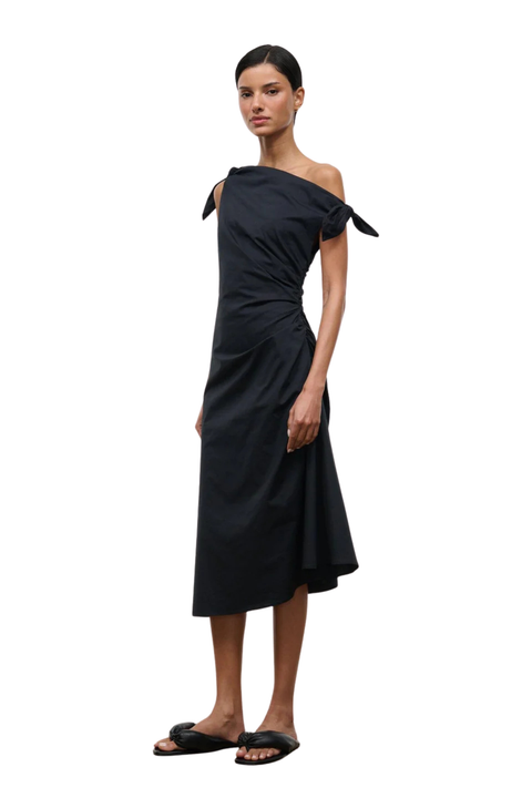A model wearing a black off-the-shoulder midi dress with tie details on the shoulders, paired with black slide sandals, set against a plain white background.