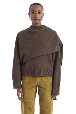A model wearing a brown asymmetrical draped knit sweater and mustard yellow pants, photographed against a solid white background.