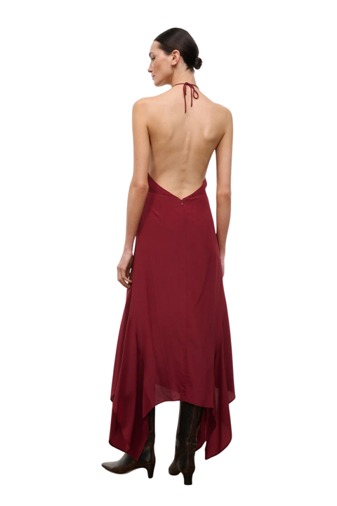 A woman shown from the back wearing a long open-back dress in deep burgundy with a tie at the neck and dark brown ankle boots, photographed against a plain white background.