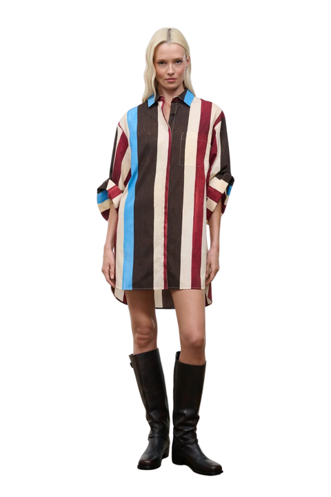 A blonde model stands against a white background wearing a long-sleeve striped shirt dress with vertical bands of cream, dark brown, red, and light blue, paired with black knee-high boots.