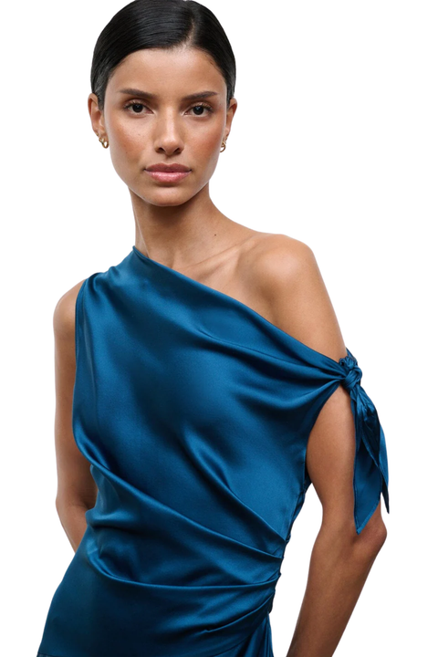 A woman modeling a satin one-shoulder dress in a rich teal blue color, featuring a side tie detail, set against a plain white background.