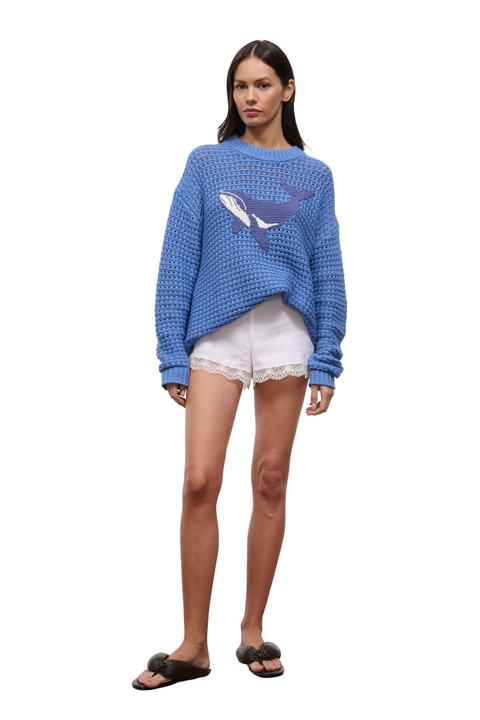 A woman modeling a blue textured knit sweater with a whale graphic on the front, paired with white shorts and black slide sandals, against a plain white background.