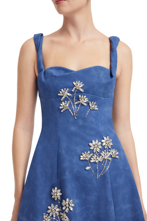 A woman wearing a blue sleeveless dress with floral embellishments on the bodice and skirt, shown against a plain white background.