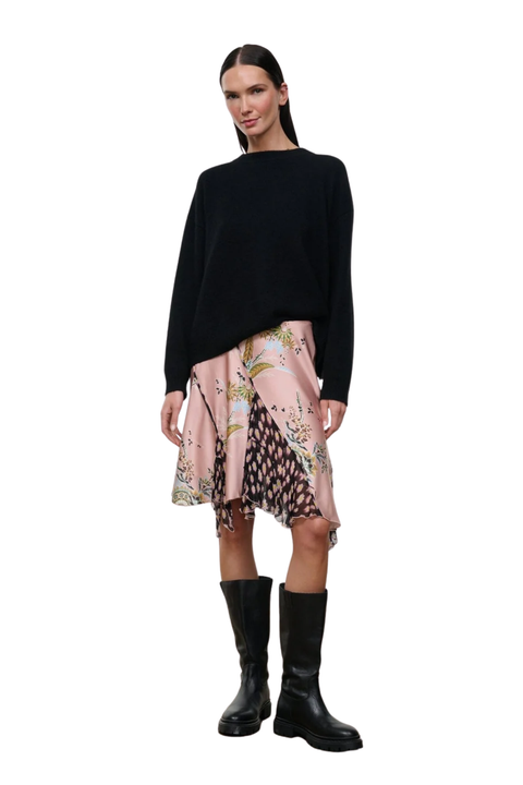 A model stands against a white background wearing a black long-sleeve knit top, a pink patterned asymmetrical skirt, and black knee-high boots.