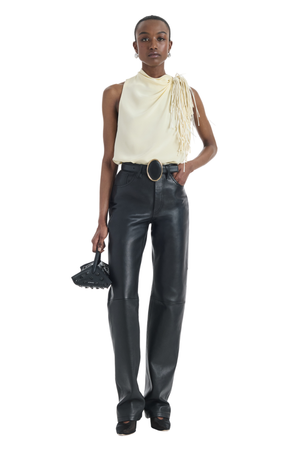 A model stands against a white background wearing a cream asymmetrical fringed top, black leather pants, a black belt with a silver oval buckle, and holding a small black textured handbag.
