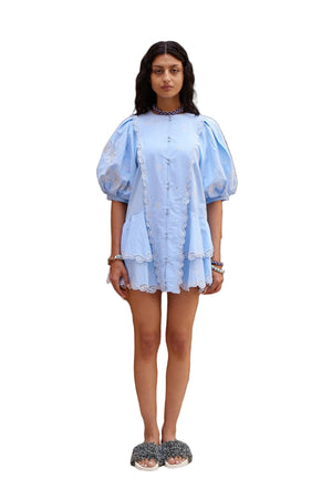 Woman wearing a light blue dress standing in a decorative outdoor setting with plants and a pool.