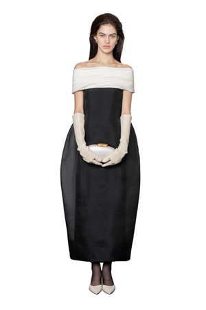 A model stands against a white background wearing a black off-the-shoulder midi dress with a wide cream band across the upper chest, cream elbow-length gloves, and holding a small cream clutch.