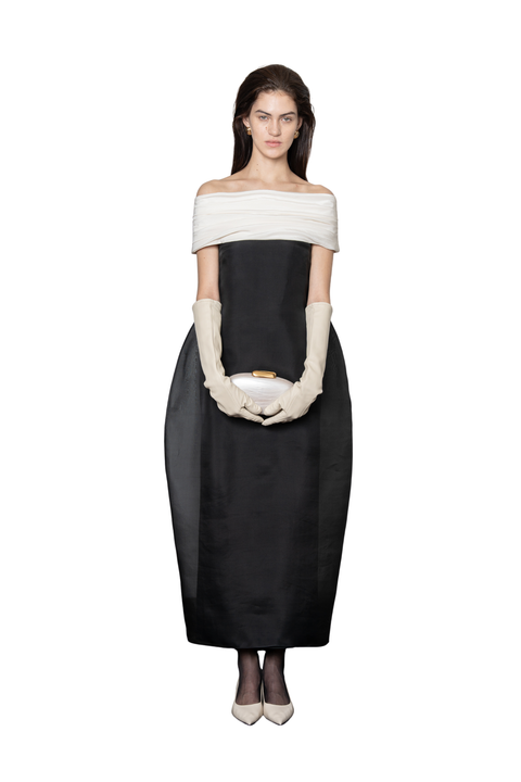 A model stands against a white background wearing a black off-the-shoulder midi dress with a wide cream band across the upper chest, cream elbow-length gloves, and holding a small cream clutch.