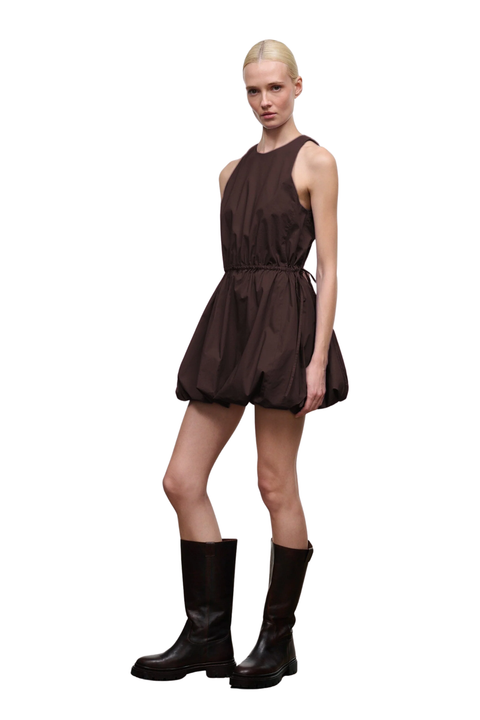 A model wearing a dark brown sleeveless mini dress with a gathered, balloon-like skirt and black knee-high boots, photographed against a solid white background.