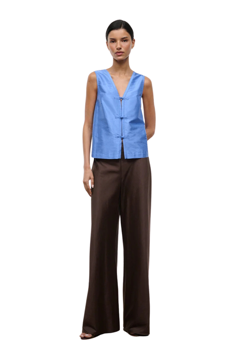 A model wearing a sleeveless light blue vest with a button closure and dark brown wide-leg trousers, photographed against a solid white background.