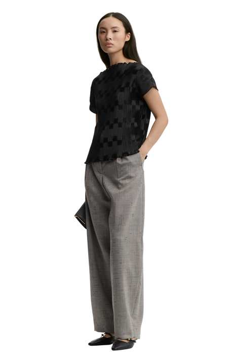 A woman modeling a black short-sleeve top with a subtle geometric pattern and wide-leg gray trousers, standing against a plain white background.