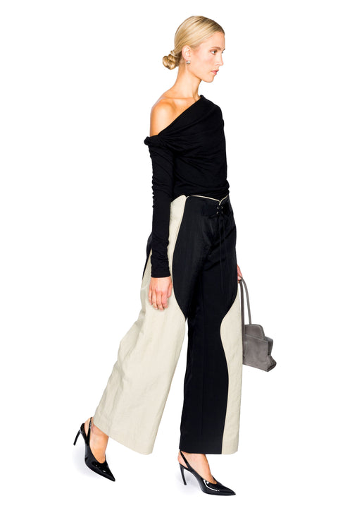 Woman facing the right wearing a black off-shoulder top and wide-leg pants with a gray handbag on a white background
