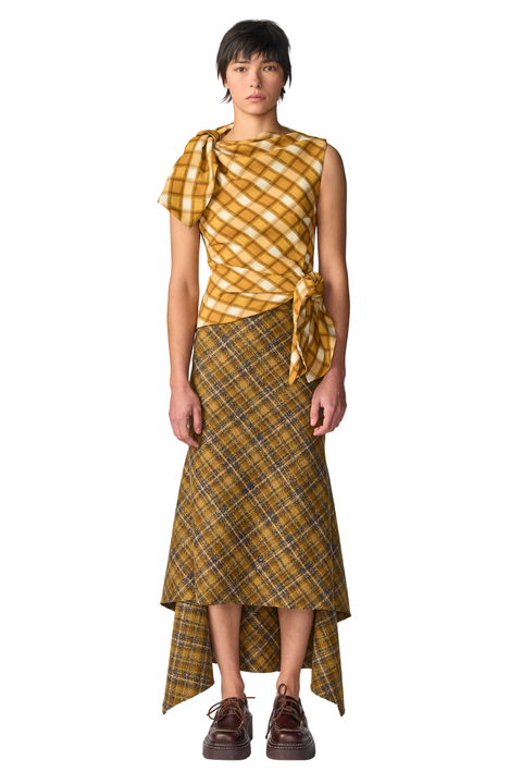 A model stands against a white studio background wearing a yellow and brown plaid outfit consisting of a sleeveless top with a tied waist and a matching high-low skirt, paired with dark brown shoes.