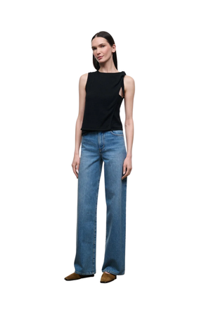 A model stands against a plain white background wearing a sleeveless black top with side cutouts, wide-leg blue jeans, and brown pointed shoes.