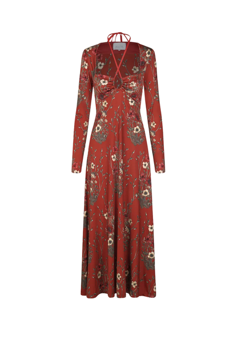 A long-sleeve maxi dress in rust red with a floral and botanical print, featuring a V-neckline with tie details at the collar and a gathered sweetheart bodice, shown against a plain white background.