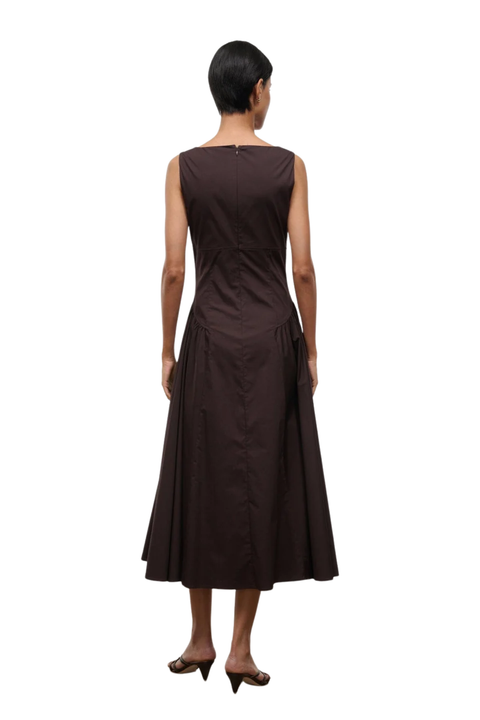 A woman viewed from the back wearing a sleeveless dark brown midi dress with a gathered waist and flared skirt, paired with dark heeled sandals, against a plain white background.