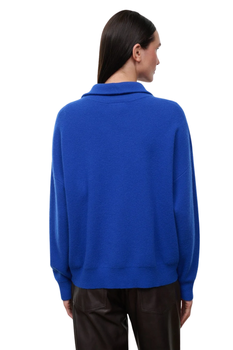 A model shown from the back wearing a bright royal blue knit sweater with a mock neck collar, paired with dark brown pants, against a plain white background.