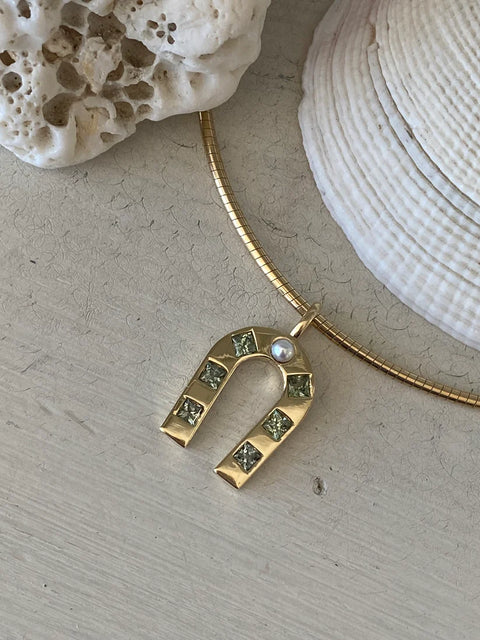 Gold horseshoe pendant with green gemstones on a gold chain, placed on a textured surface with a shell in the background.