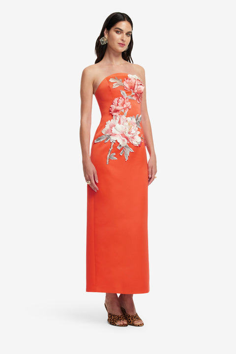 Woman posing in an orange strapless maxi length dress with floral patterns on a white background