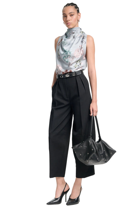 Woman wearing a bias cut sleeveless floral top and black pants, holding a black handbag on a white background.