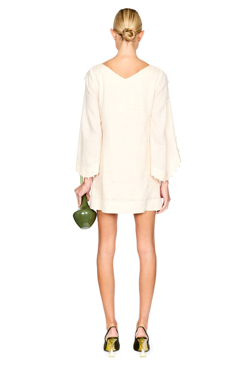 A woman shown from the back wearing a cream-colored mini dress with long bell sleeves and scalloped edges. She is holding a small green clutch and wearing metallic green and yellow shoes against a solid white background.