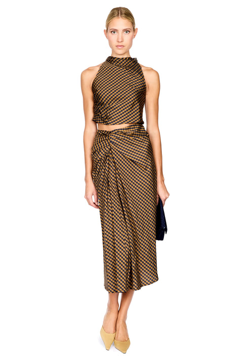A model stands against a white background wearing a brown and navy checkered halter crop top and matching draped midi skirt, paired with beige pointed-toe heels and holding a black clutch.