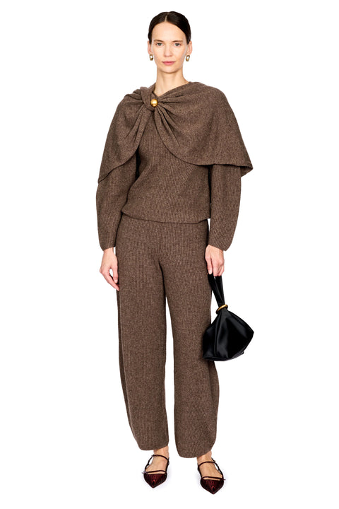 A model wearing a brown knit matching set consisting of a long-sleeve top with a draped collar and broch, and matching trousers, styled with dark pointed flats and a black handbag against a white background.